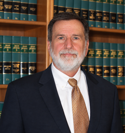 Man in suit and tie, smiling, wearing glasses, in front of a bookcase.