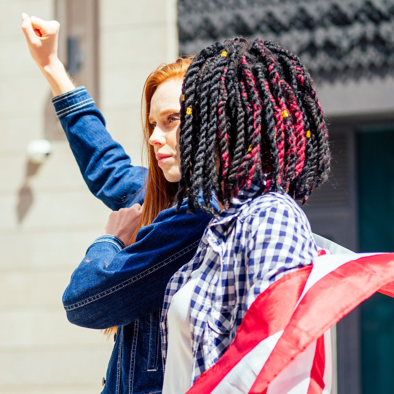 Two people at a protest: one raises a fist, the other holds a US flag. Both wear denim and look forward.