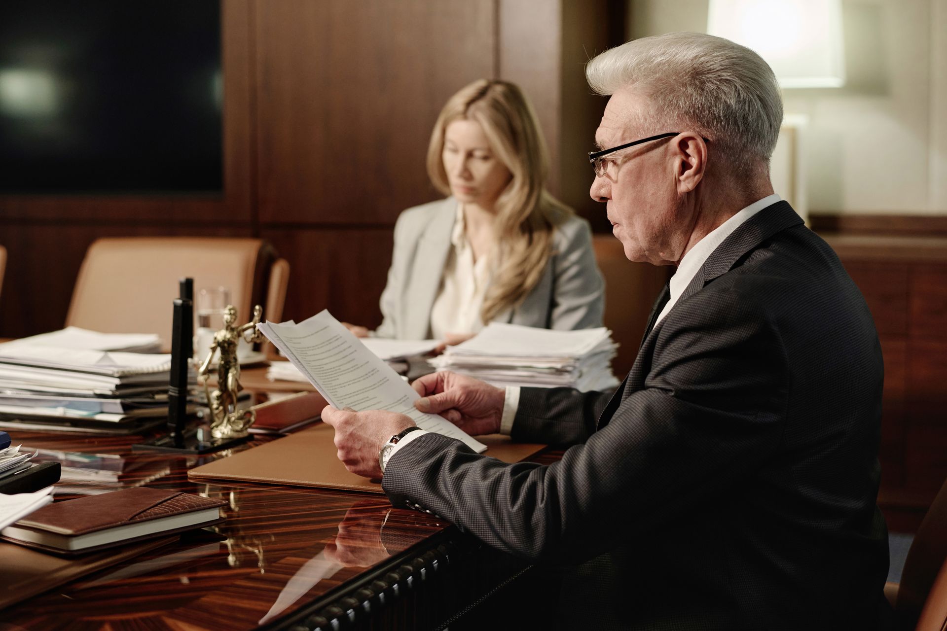 Lawyer in suit reads document at table, another person in background.