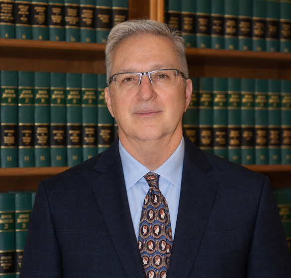 Man in suit and glasses smiling in front of a bookshelf filled with legal books.