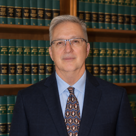 Man in suit and glasses smiling in front of a bookshelf filled with legal books.