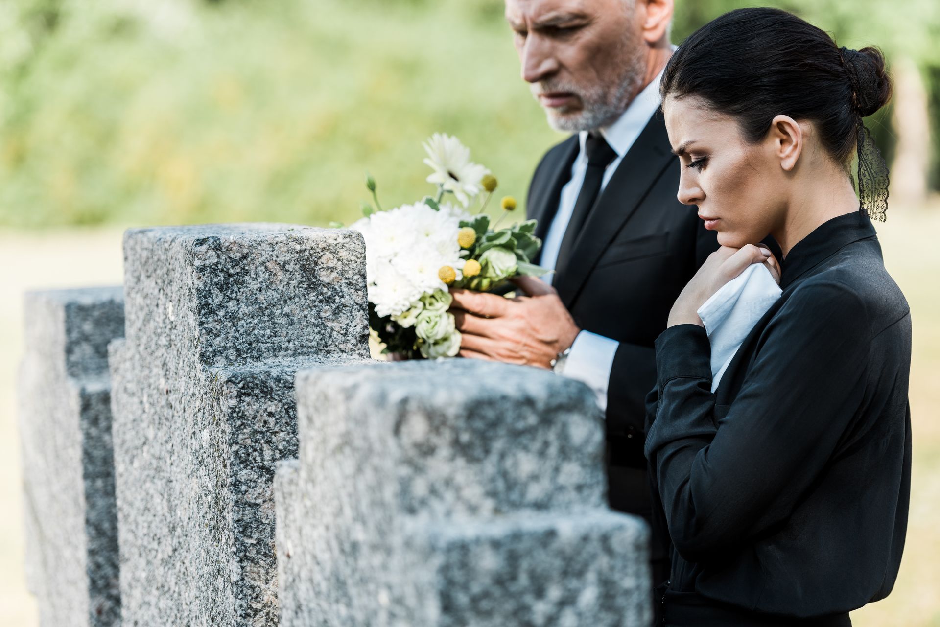 Man in suit places flowers on tombstone; woman in black grieves nearby.
