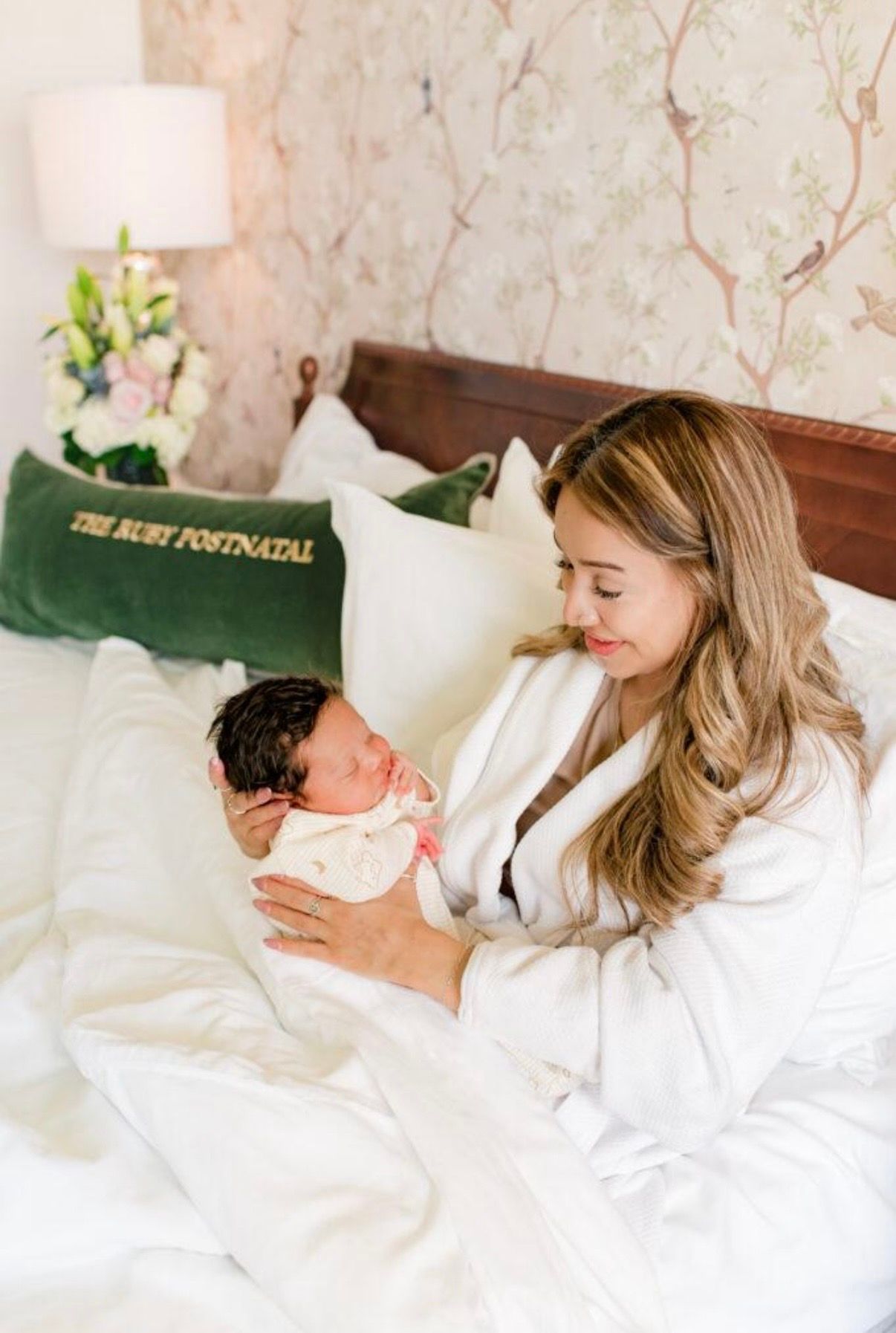Mother holding newborn in bed, smiling in a bright bedroom with white bedding and green pillows