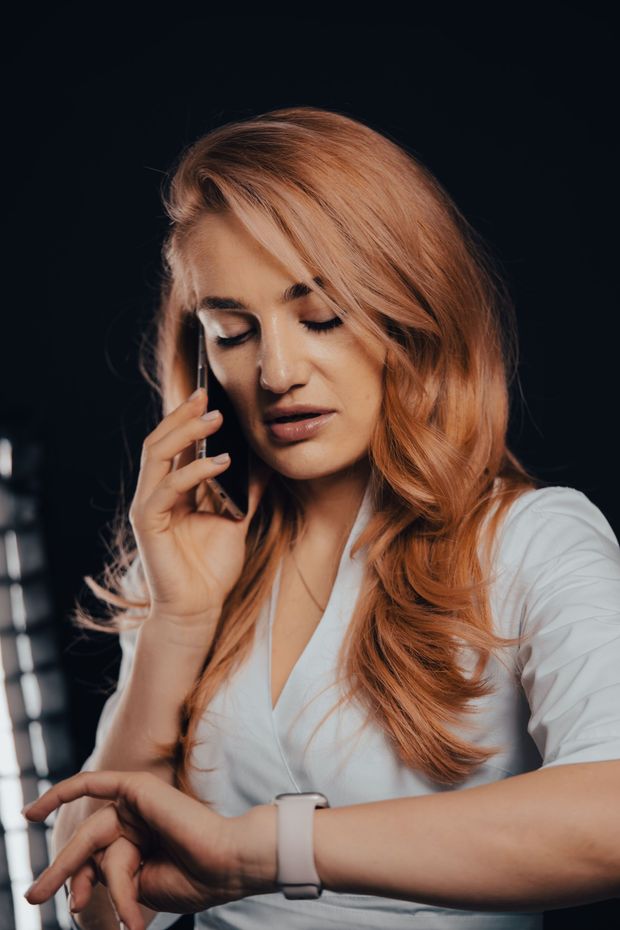 Woman with long red hair looks at her smartwatch while holding a phone on a dark background