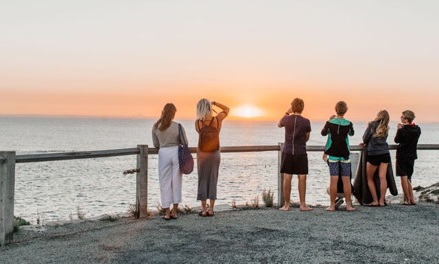 Group of people standing by a waterfront fence at sunset, taking photos of the orange horizon.