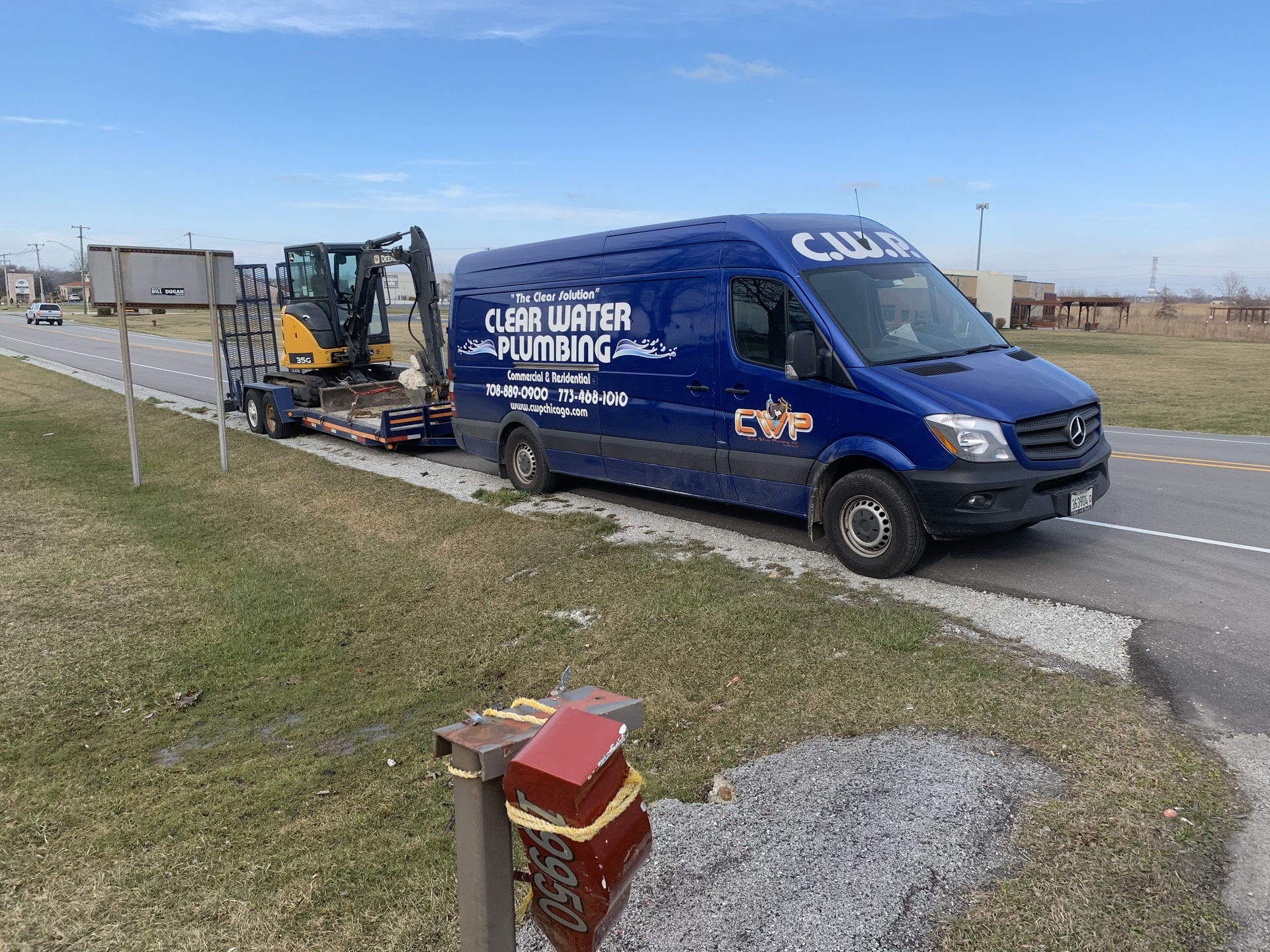Blue roadside work van with trailer and mini excavator parked on a highway shoulder beside a fire hydrant.