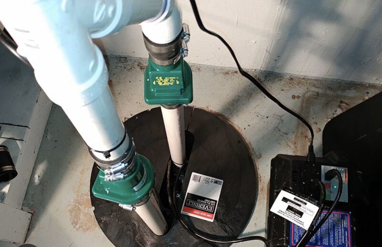 Two blue pipes with green pumps over a round floor opening next to a wall and cables