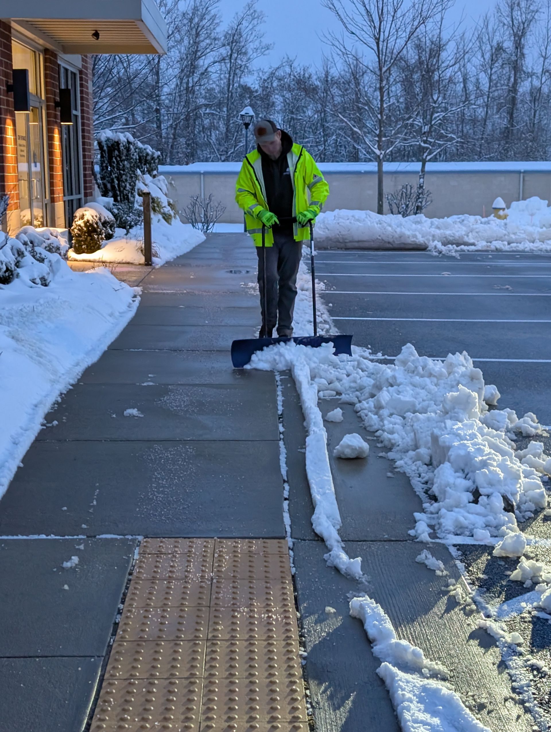 A man in a yellow jacket is shoveling snow on a sidewalk
