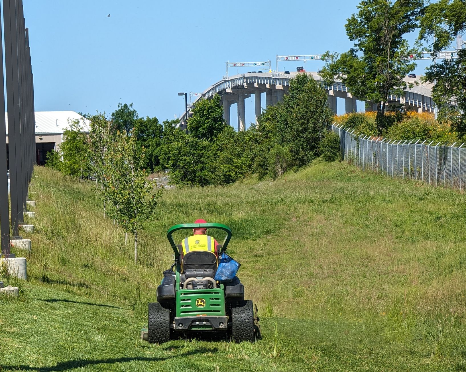 A person is cutting grass with a riding lawn mower next to a freeway.