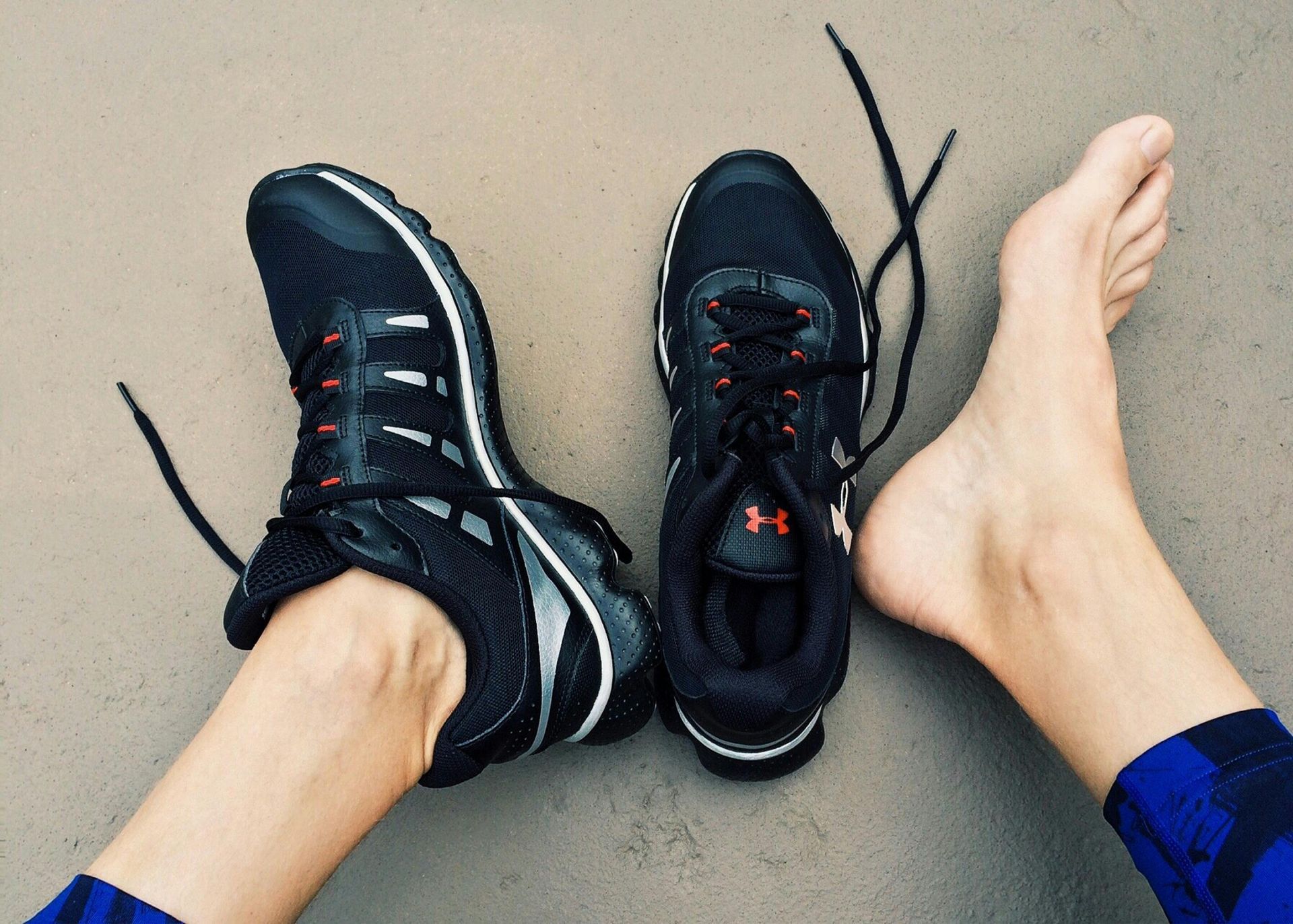 Black athletic shoes next to a bare foot on a neutral-toned surface.