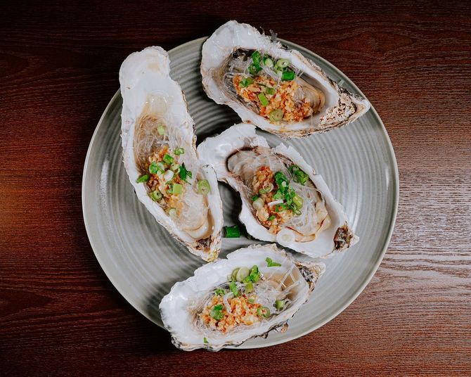 Four oysters on a gray plate with chopped herbs and sauce, on a dark wooden table