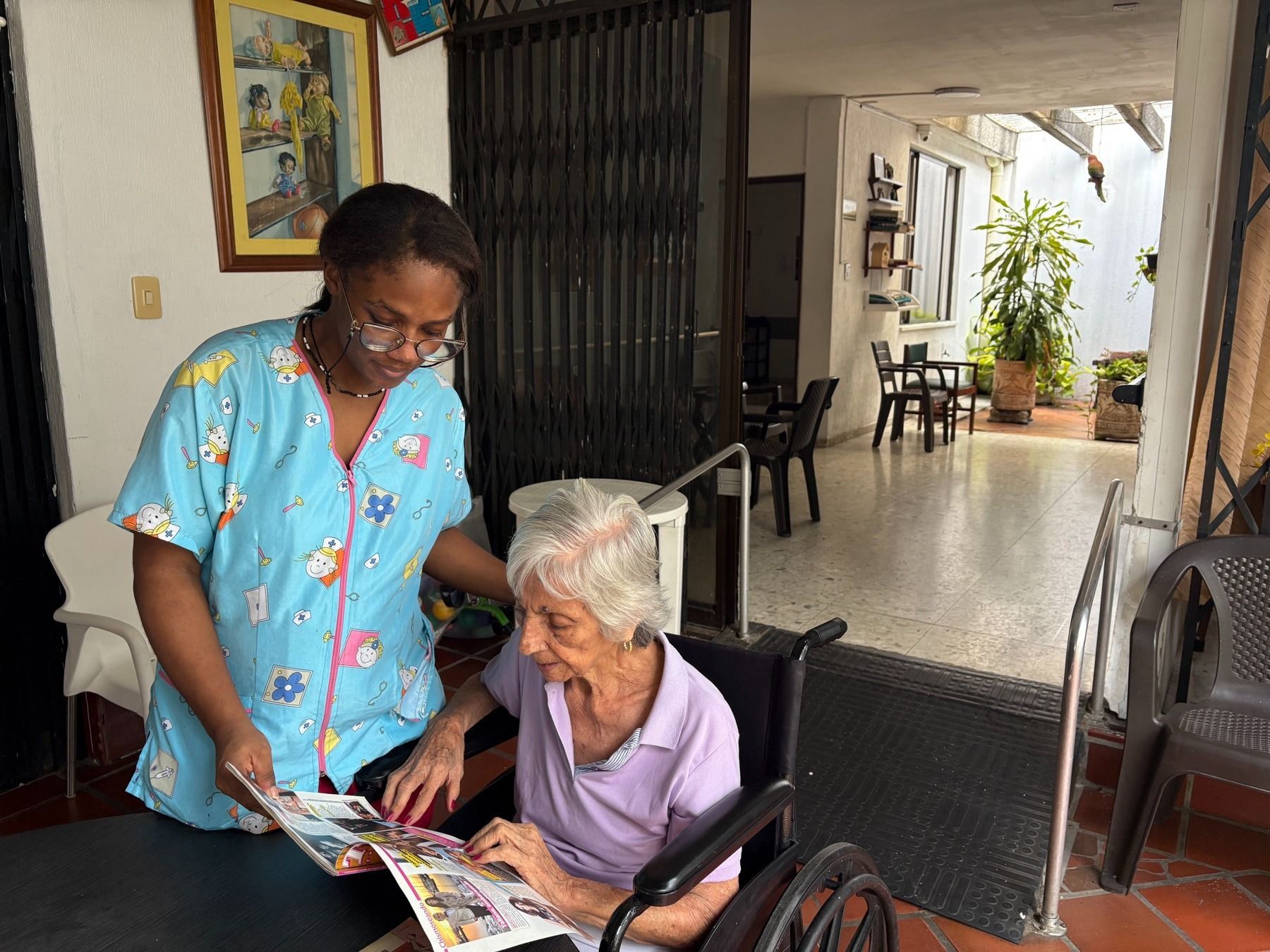 Una cuidadora le muestra una revista a una mujer mayor sentada en una silla en un porche cubierto
