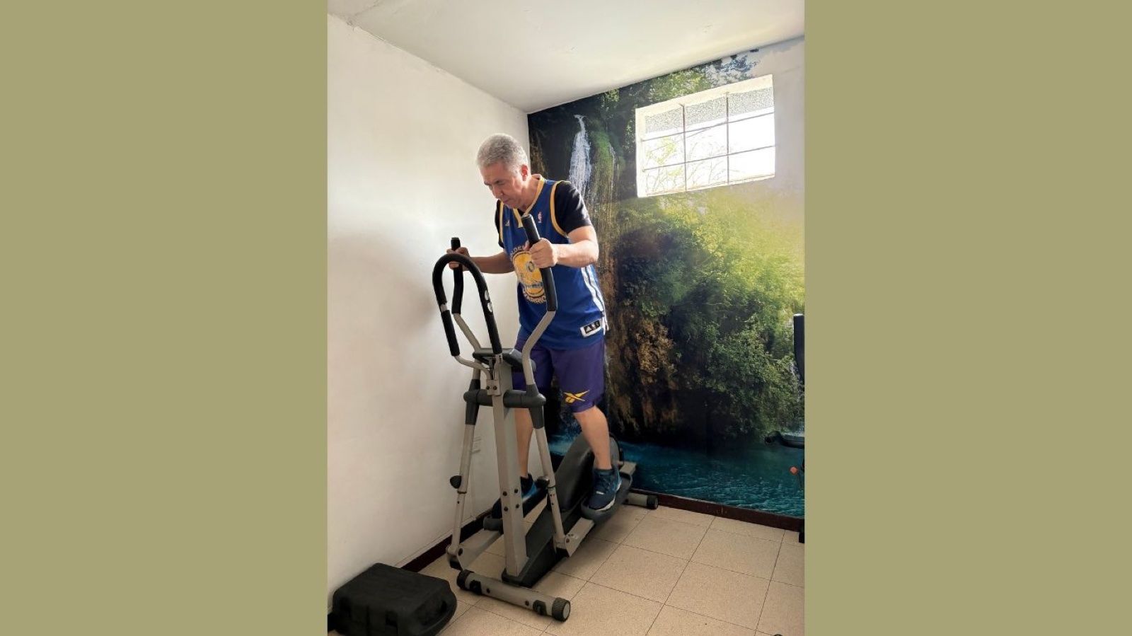 Una persona haciendo ejercicio en una máquina elíptica en una habitación pequeña con un mural en la pared y una ventana.