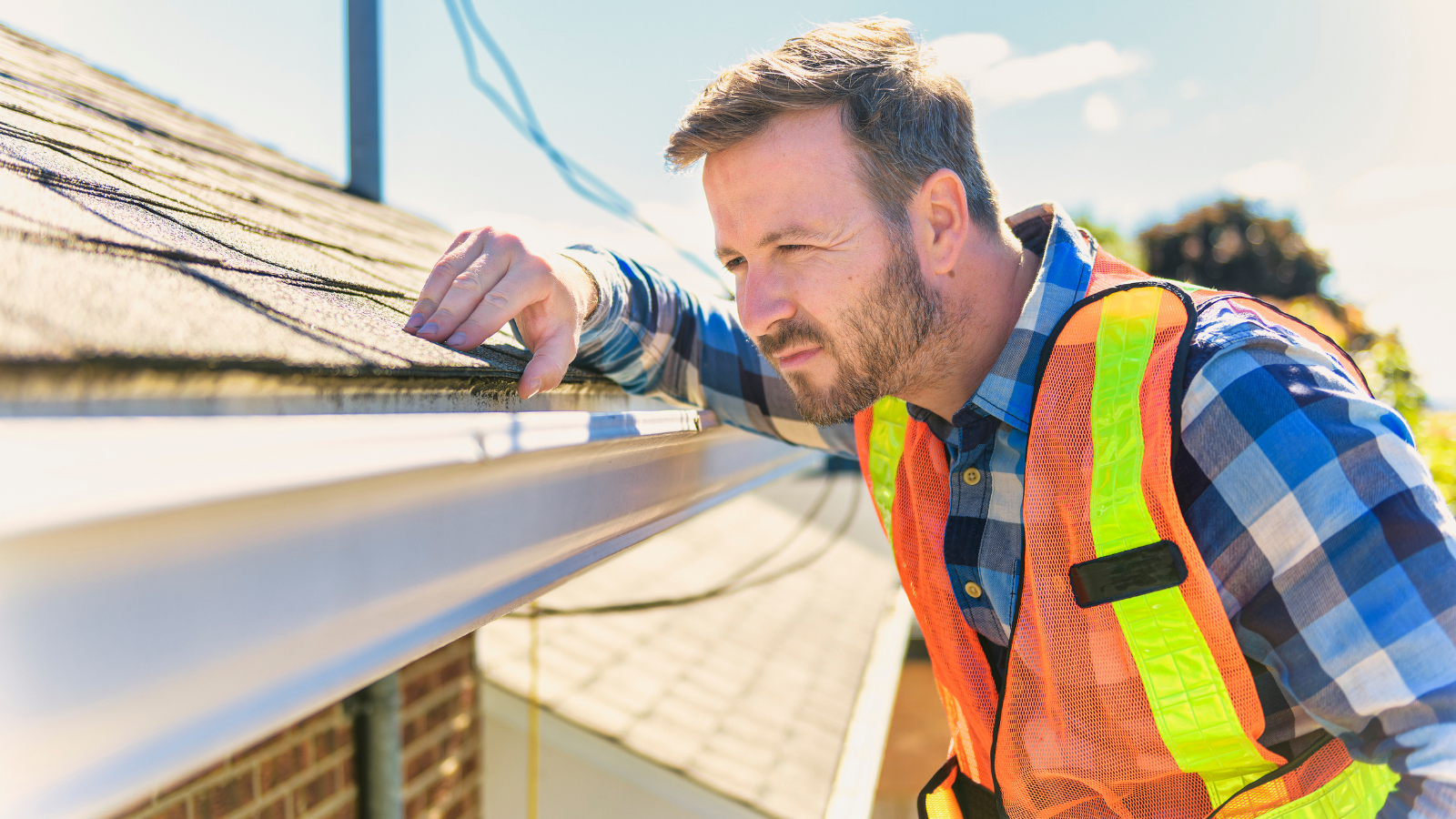 Man in safety vest inspecting a gutter on a roof, sunny day.