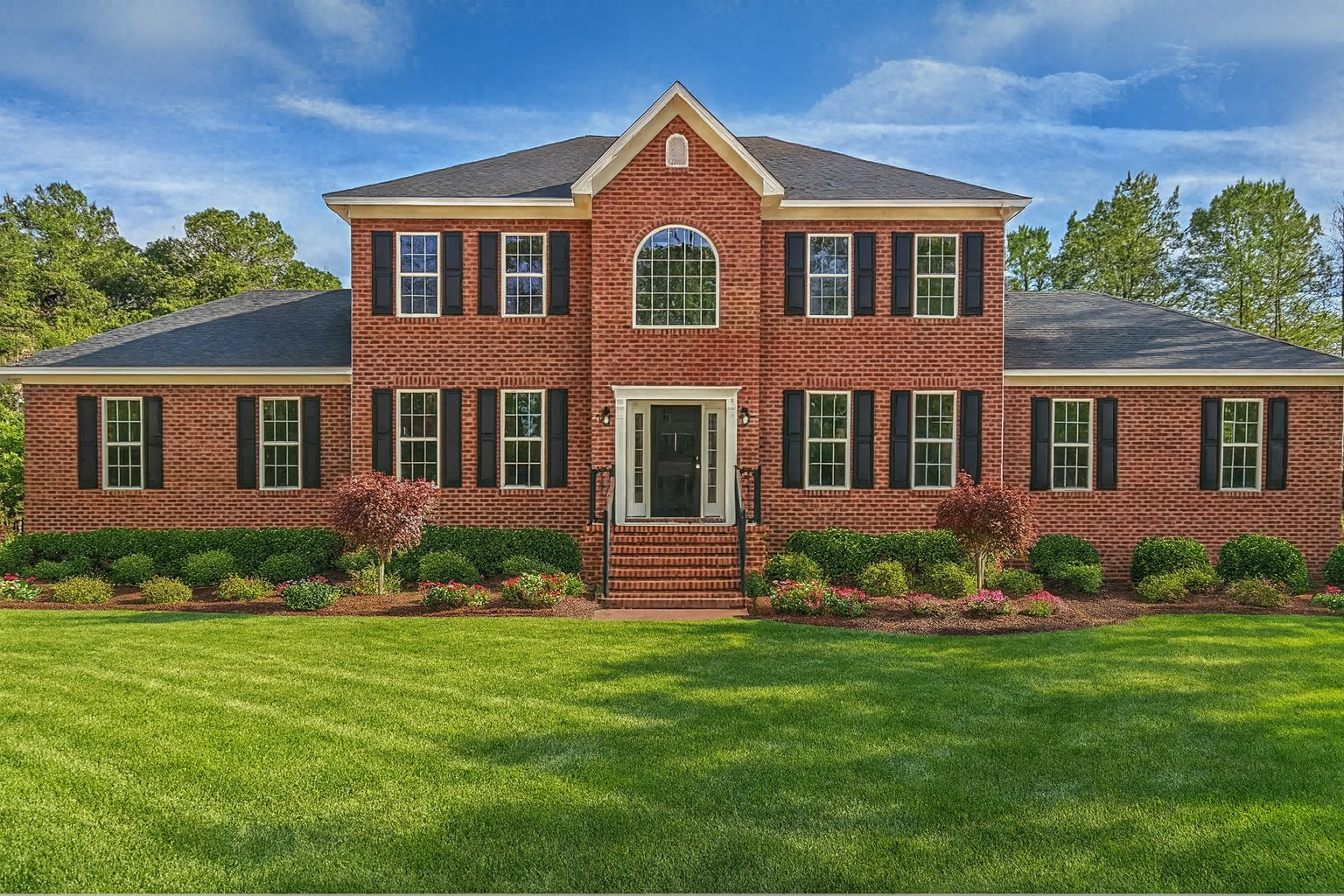 Two-story brick house with black shutters, green lawn, and landscaping under a blue sky.