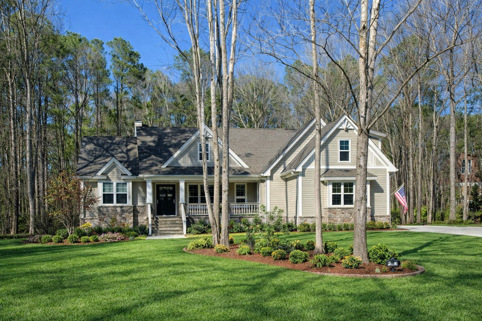 Tan house with stone accents, a porch, and American flag on a green lawn surrounded by trees.