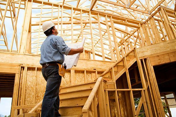 Construction worker in hard hat inspecting a wooden house frame, holding blueprints.