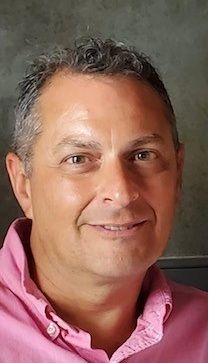 Man with short graying hair, smiling, wearing a pink button-down shirt.
