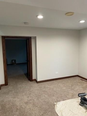 Empty beige room with carpet, white ceiling lights, and an open doorway to a dark adjacent room