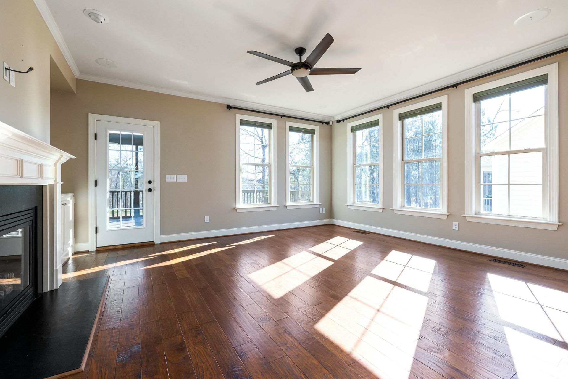 Bright empty living room with hardwood floors, fireplace, ceiling fan, and sunlit windows