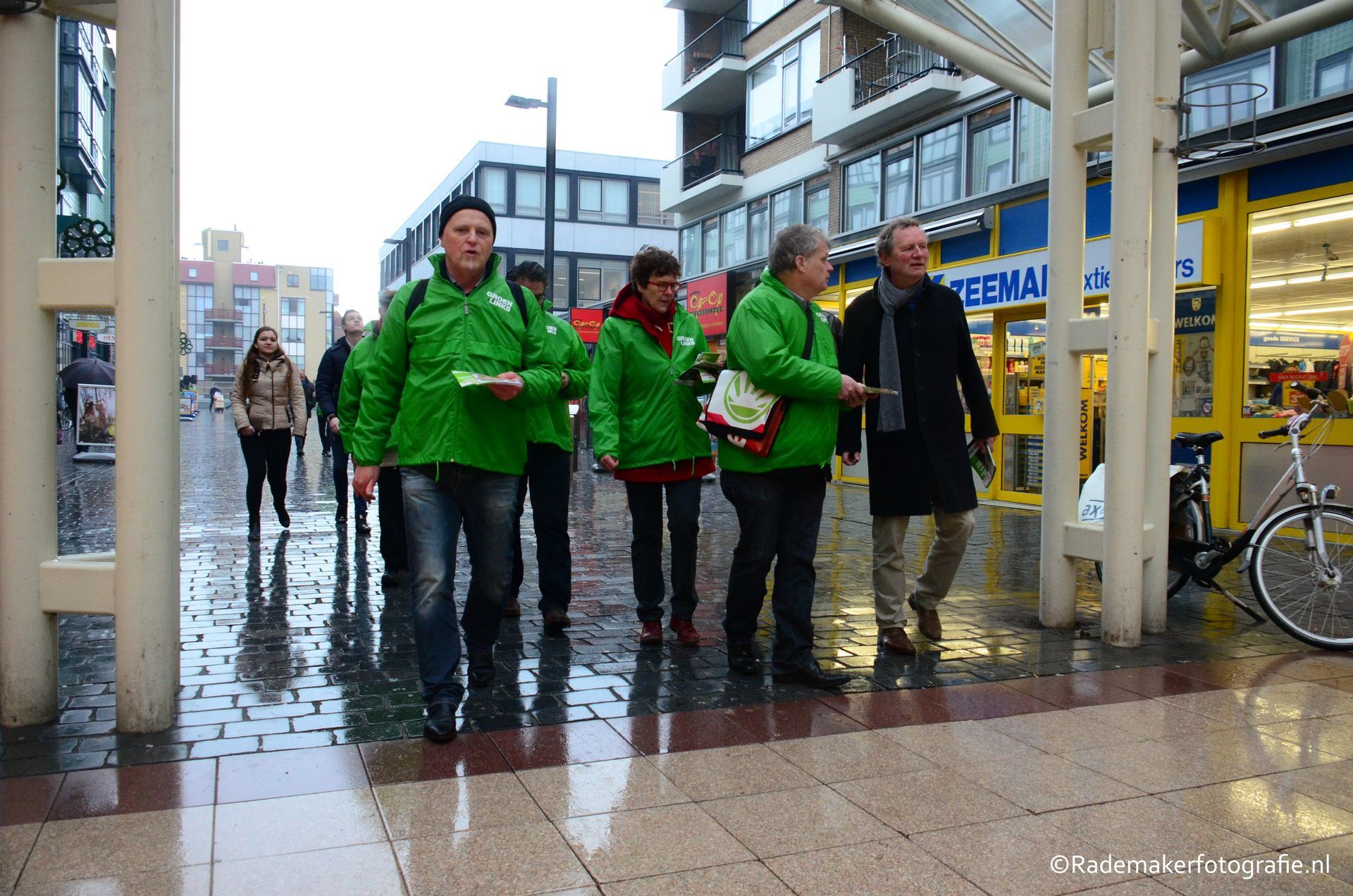 Een groep loopt door een door de regen gladde winkelstraat in de openlucht, waarvan sommigen felgroene windjacks dragen.