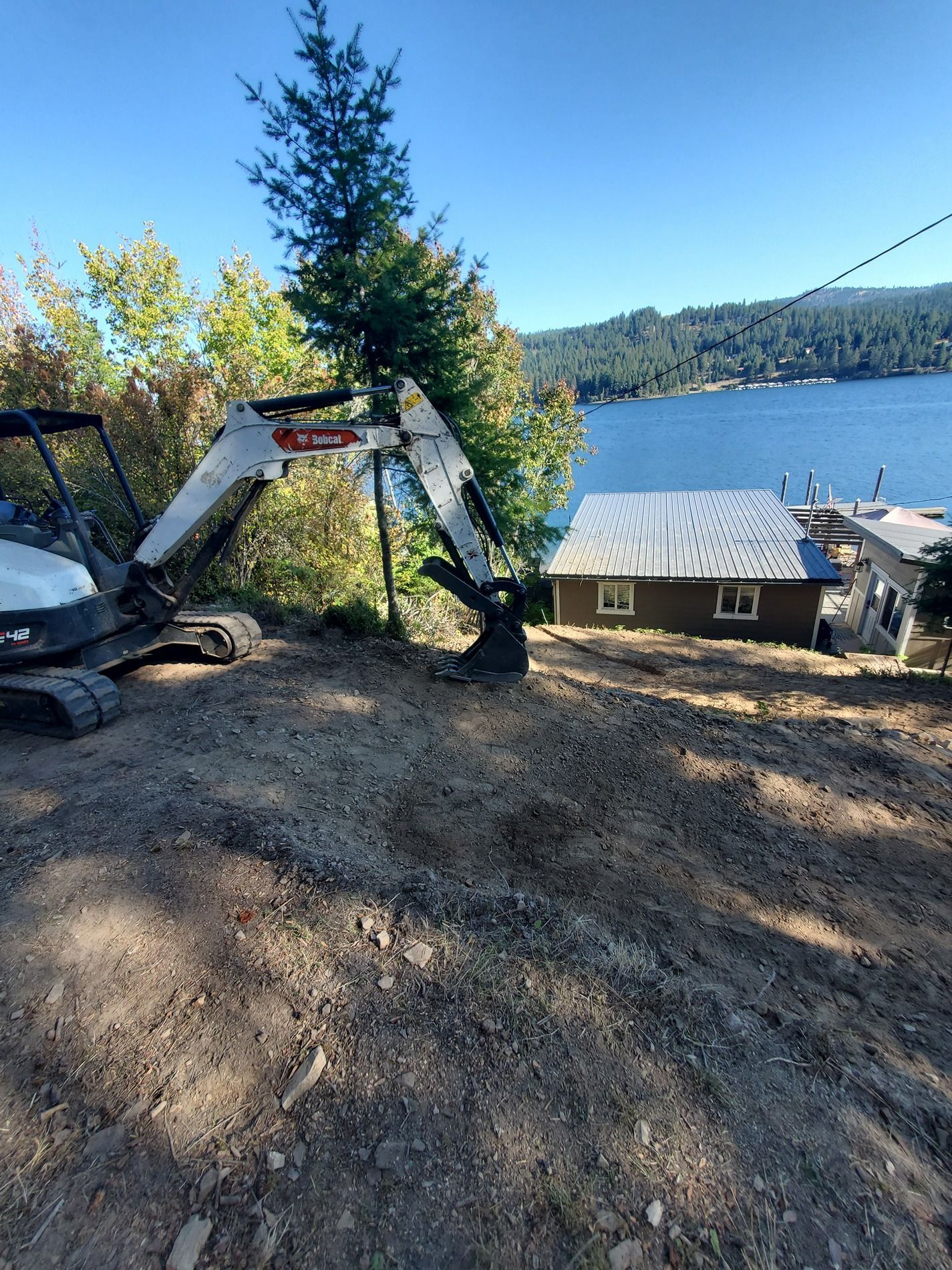 A Bobcat excavator on a dirt hillside overlooking a lake and a house with a patterned roof.