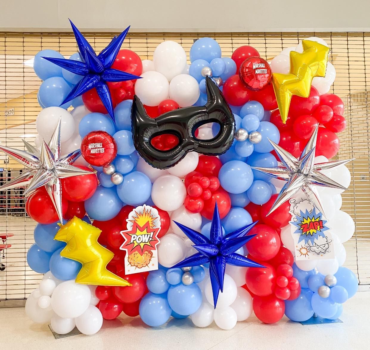 Superhero-themed balloon wall: blue, red, white balloons with star, lightning, and mask decorations.
