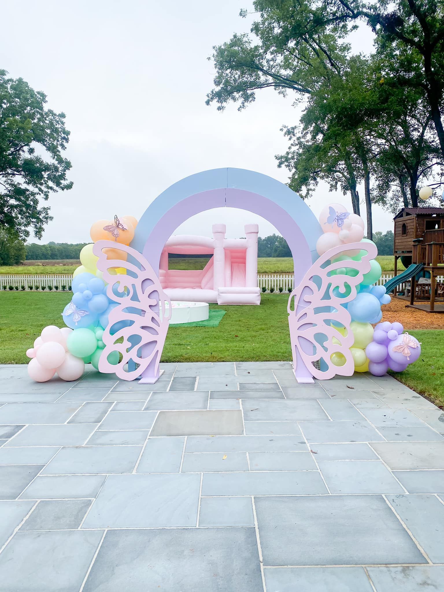 Butterfly-themed party arch with pastel balloons, a pink bounce house in the background, set on a stone patio.