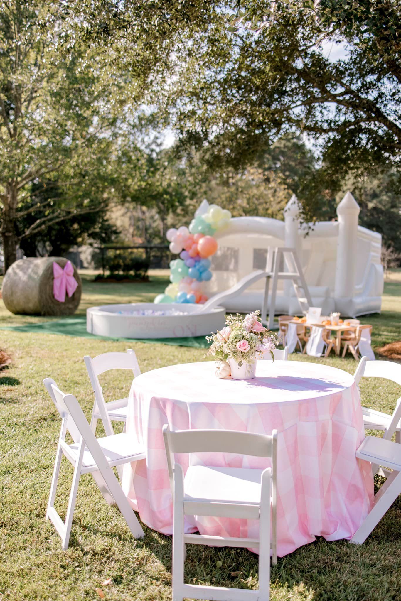 Outdoor party scene with white chairs, pink checkered table, flowers, and a white bounce house.