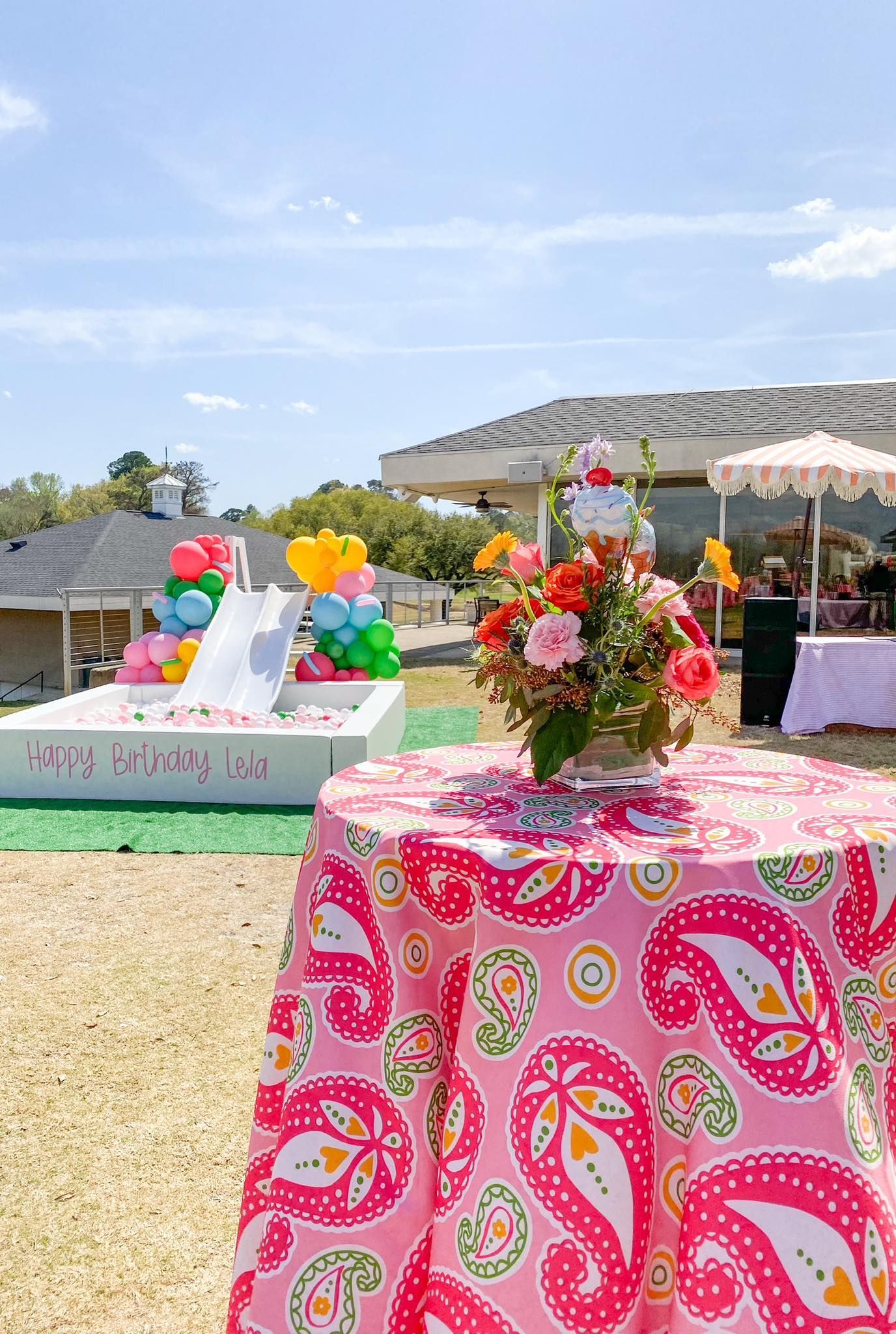 Outdoor party scene with table set for guests; pink paisley tablecloth, floral arrangement, colorful balloons, sunny sky.