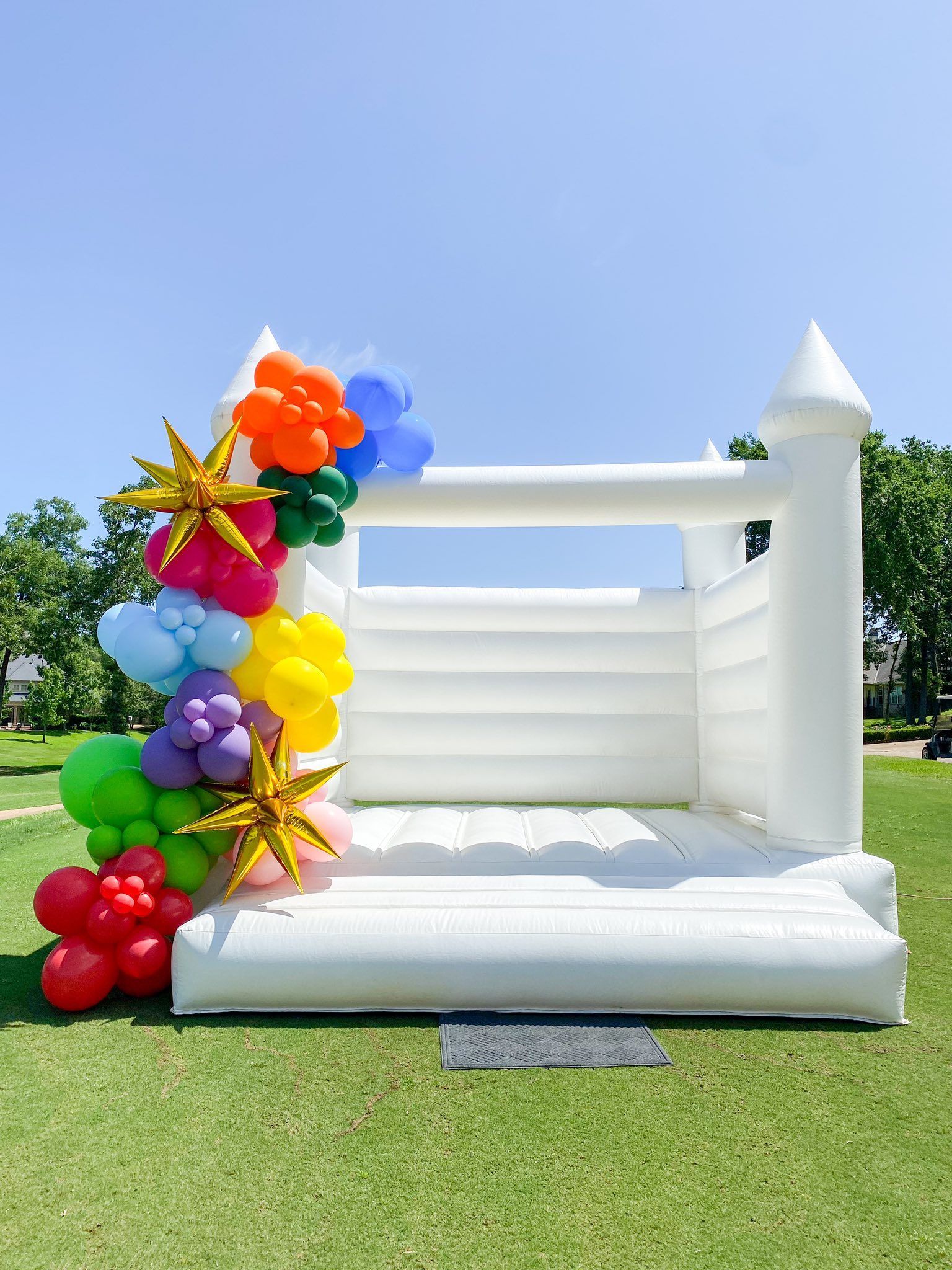 White bounce house decorated with a colorful balloon arch on a grassy lawn under a blue sky.