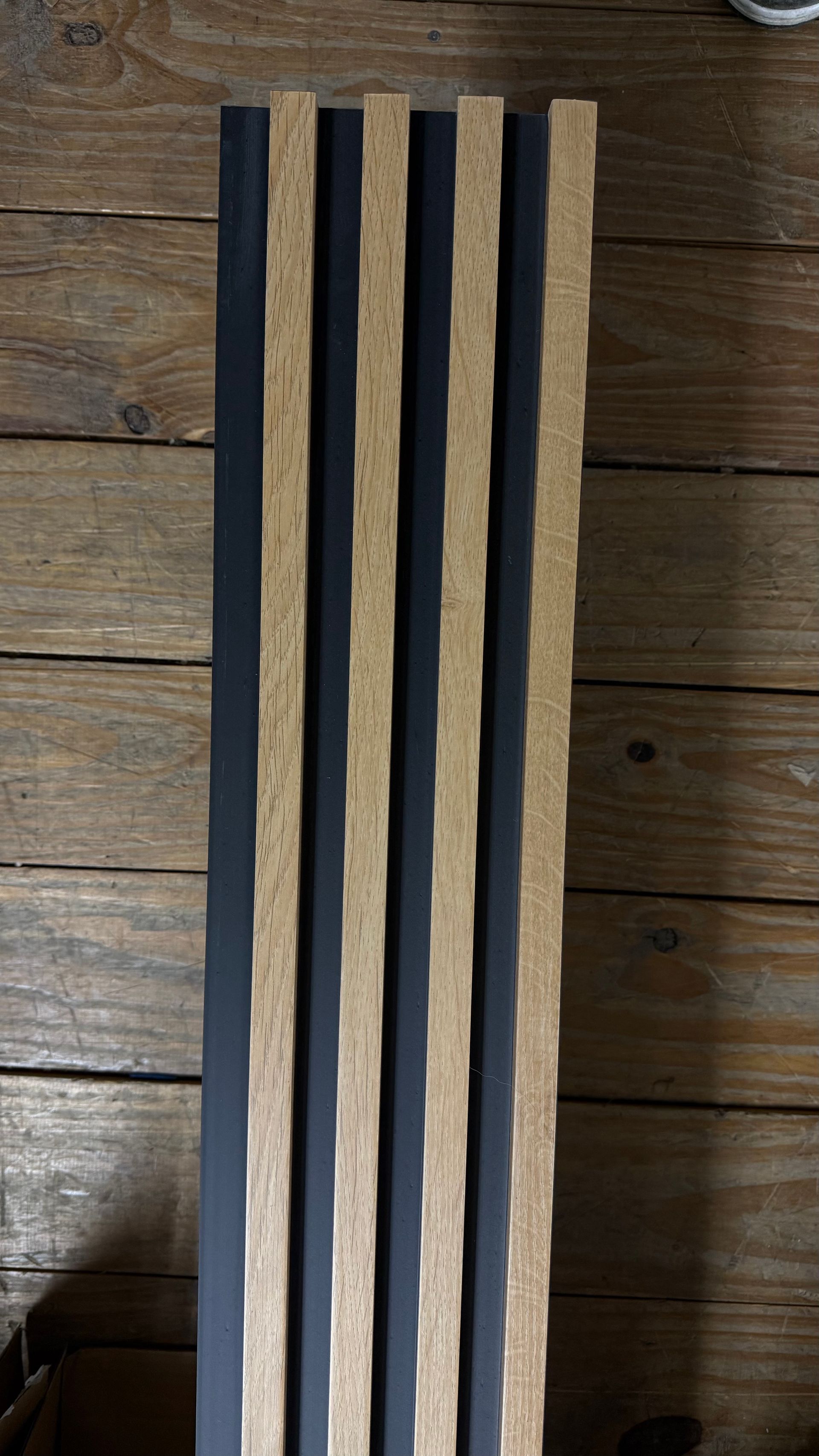 Panel vertical de listones de madera sobre un suelo de madera oscura, con franjas alternas de madera clara y negro.