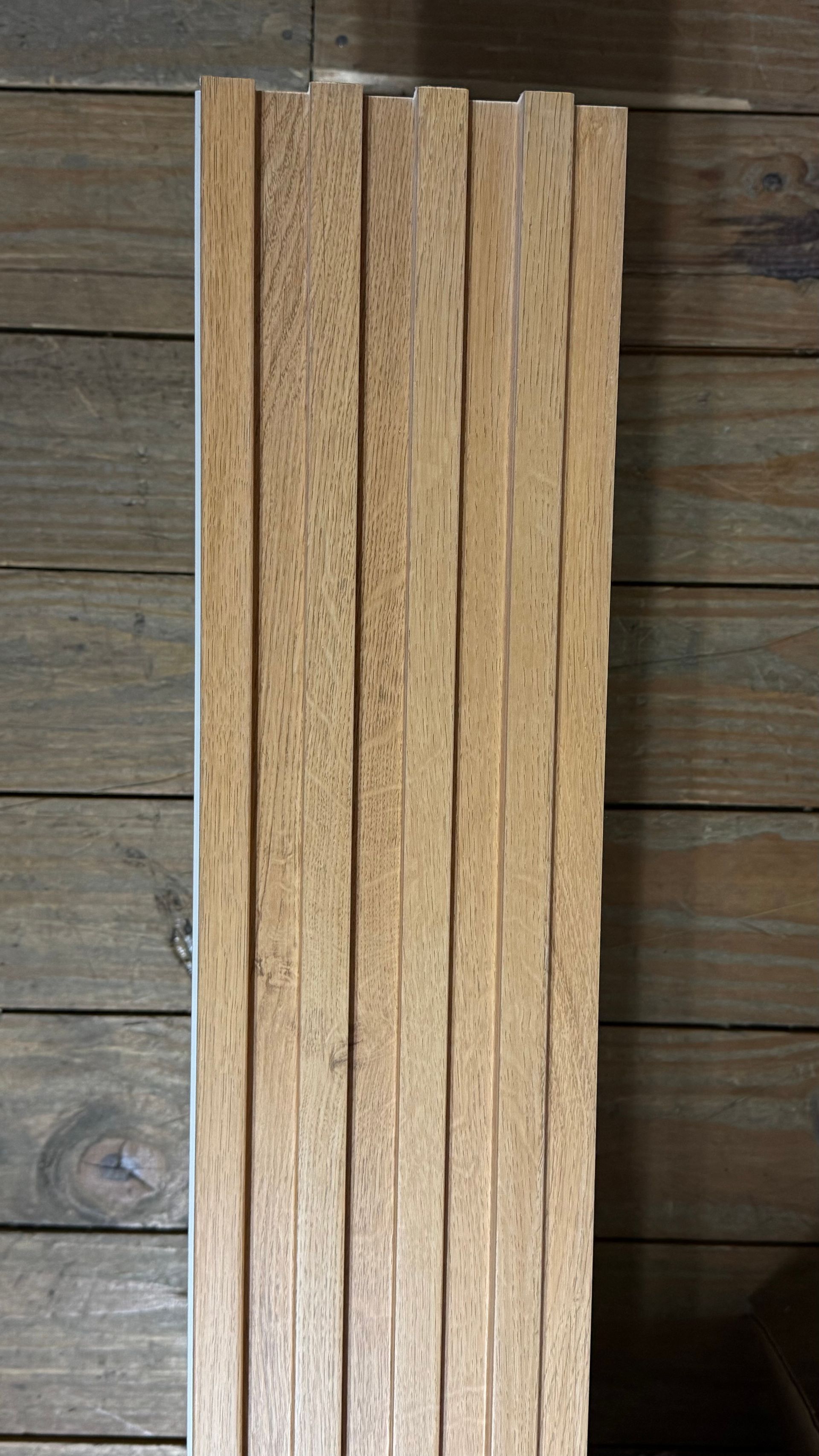 Pila de tablas estrechas de madera clara apoyadas verticalmente contra una pared de madera desgastada.