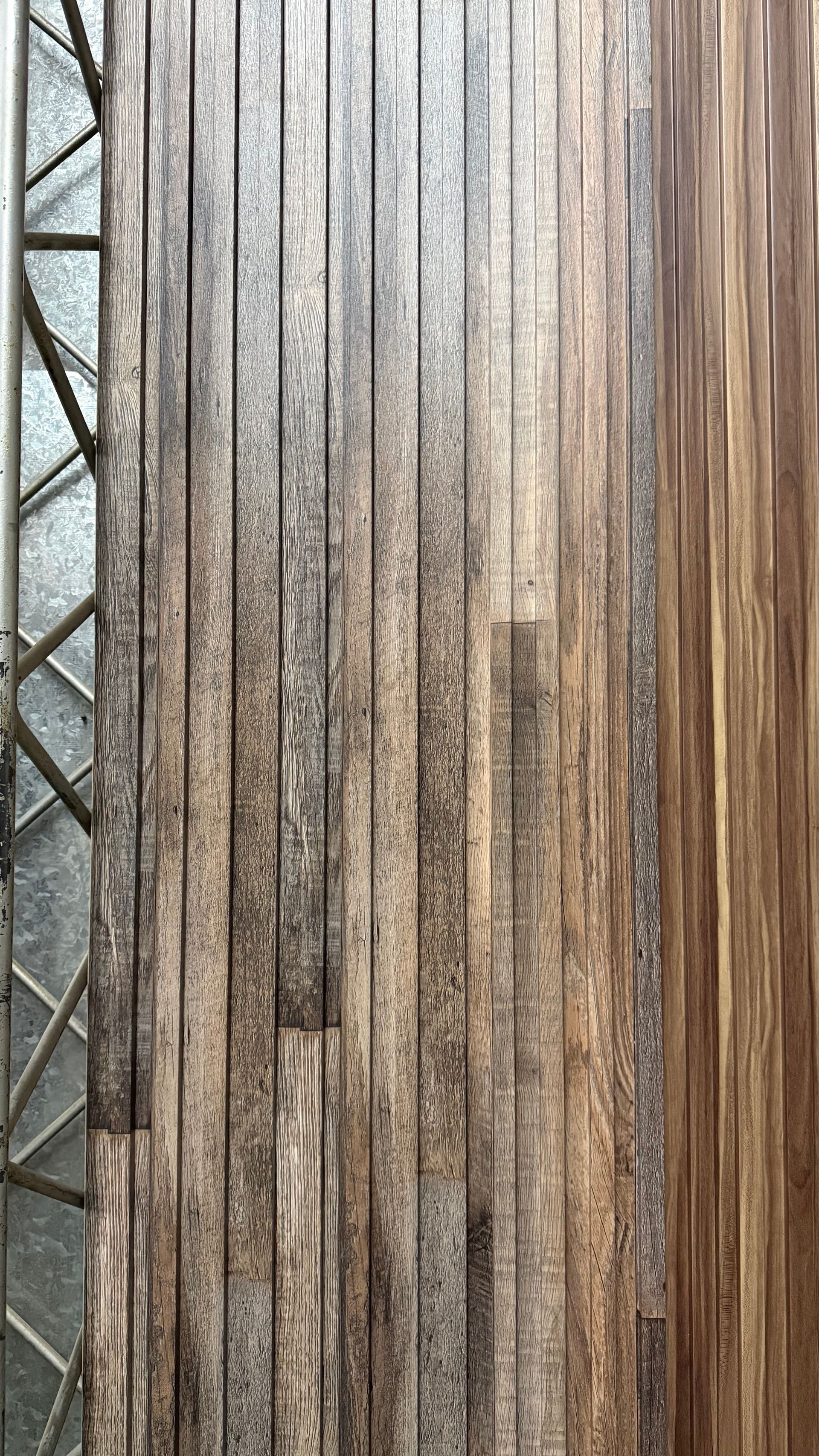 Listones de madera verticales con luz solar y sombra, que dejan ver una valla o pantalla texturizada.