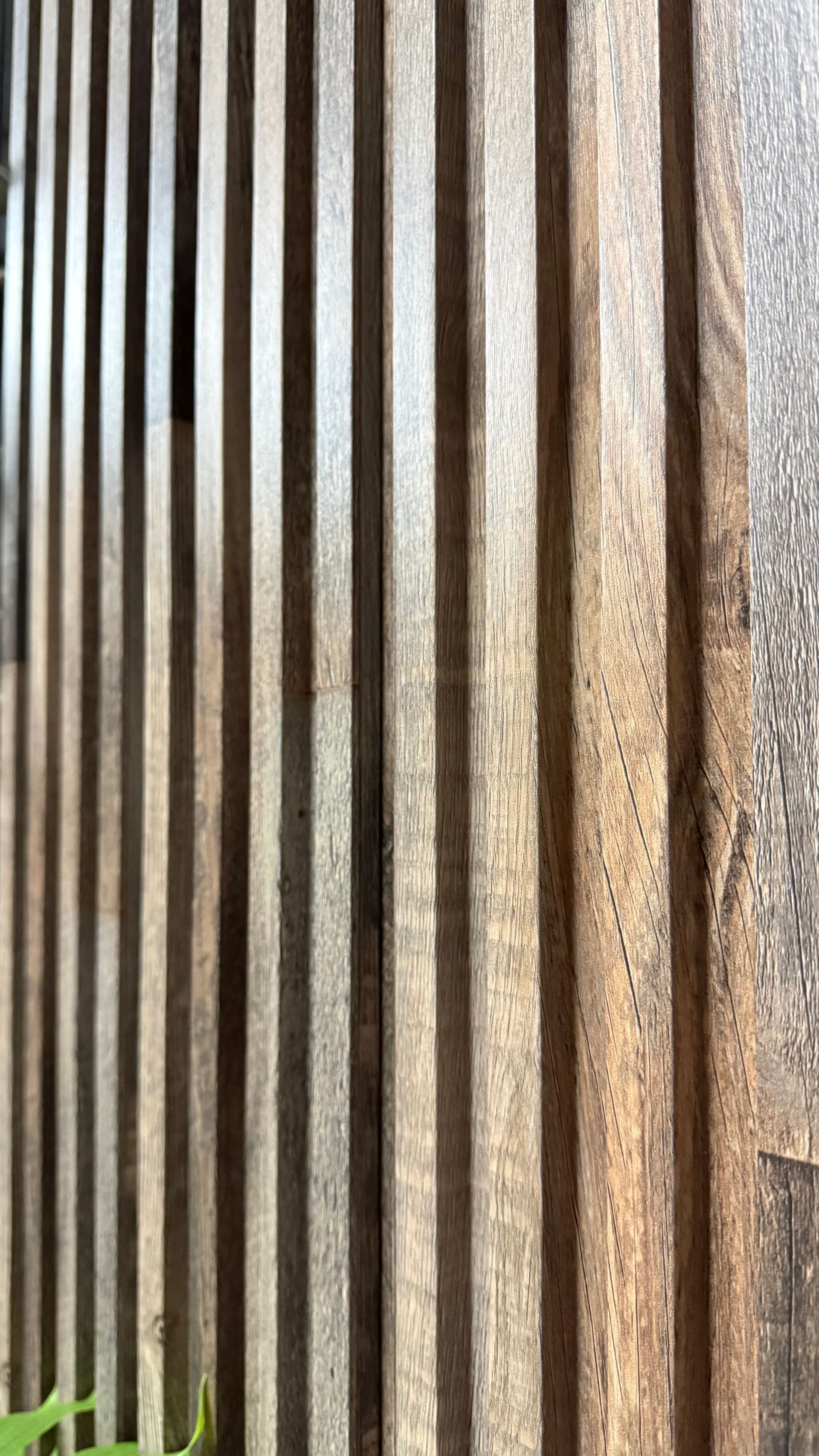 Listones de madera verticales con luz solar y sombra, que dejan ver una valla o pantalla texturizada.