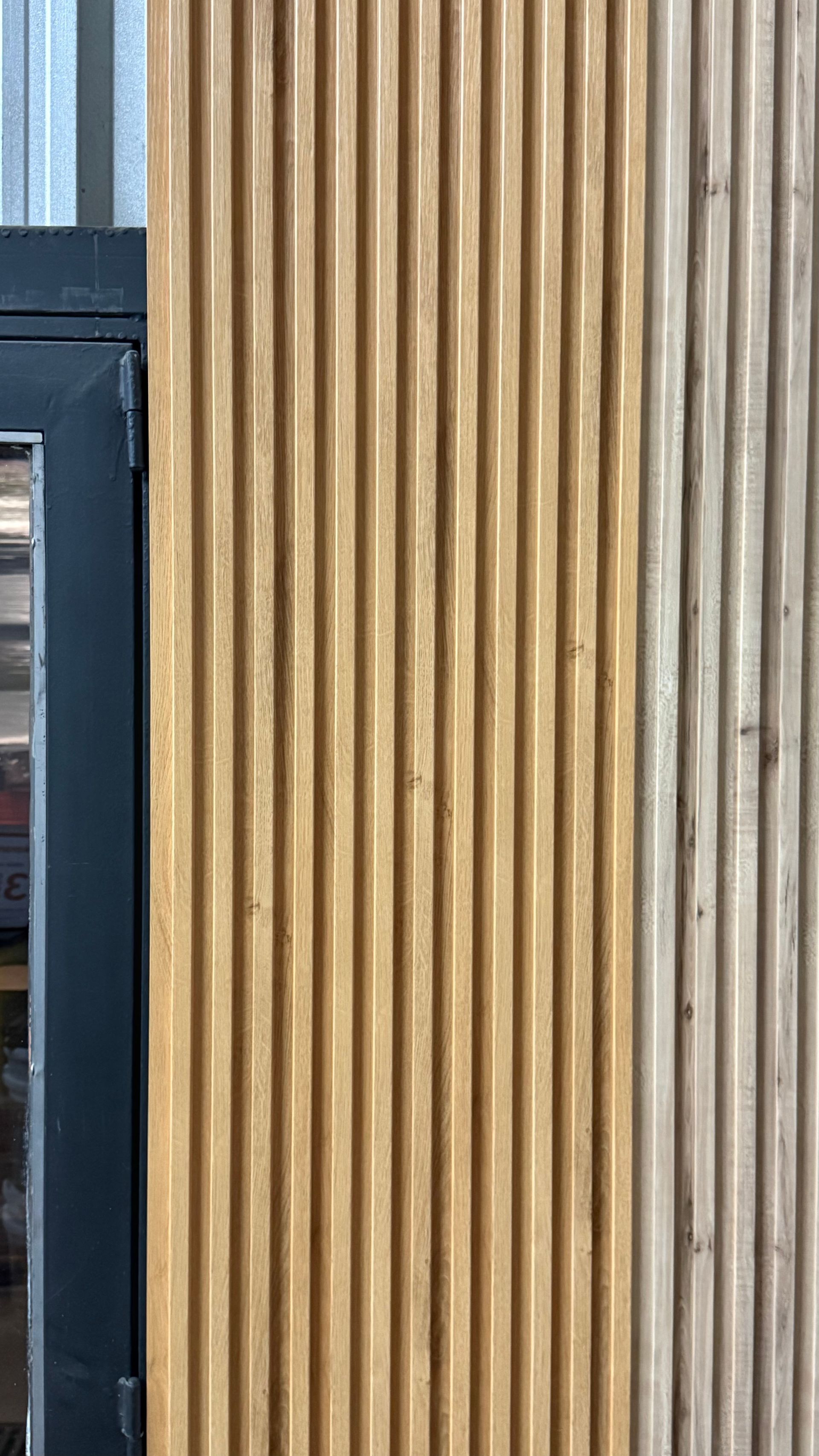 Pila de tablas estrechas de madera clara apoyadas verticalmente contra una pared de madera desgastada.