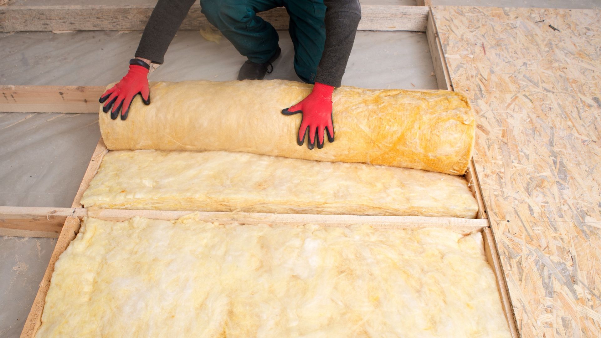 Person rolling yellow insulation batts into place in an unfinished attic, wearing red gloves.