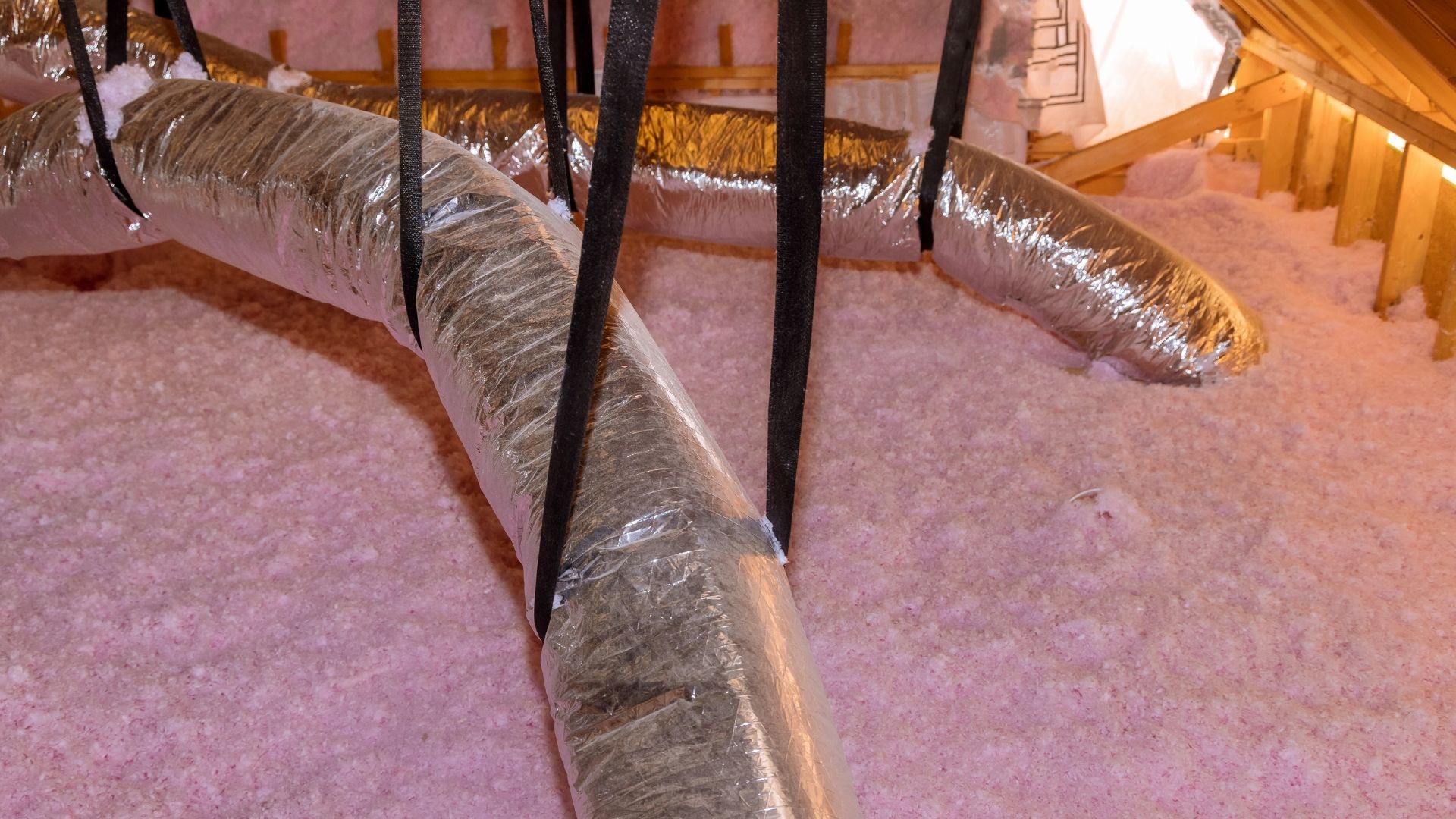 Silver insulated ductwork lying on pink insulation in an attic crawlspace.
