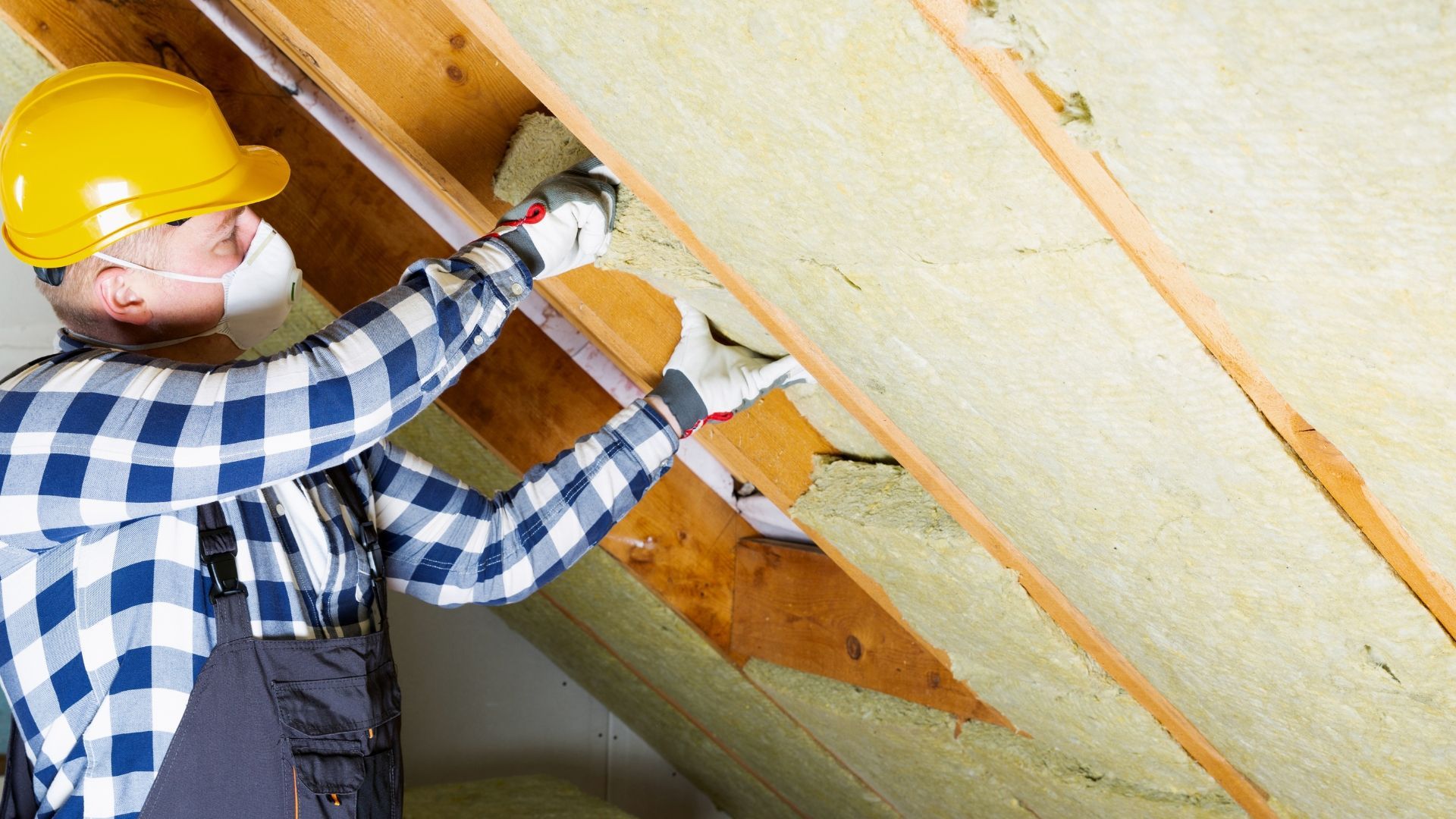 Worker in yellow hard hat installing insulation in an attic with a power drill