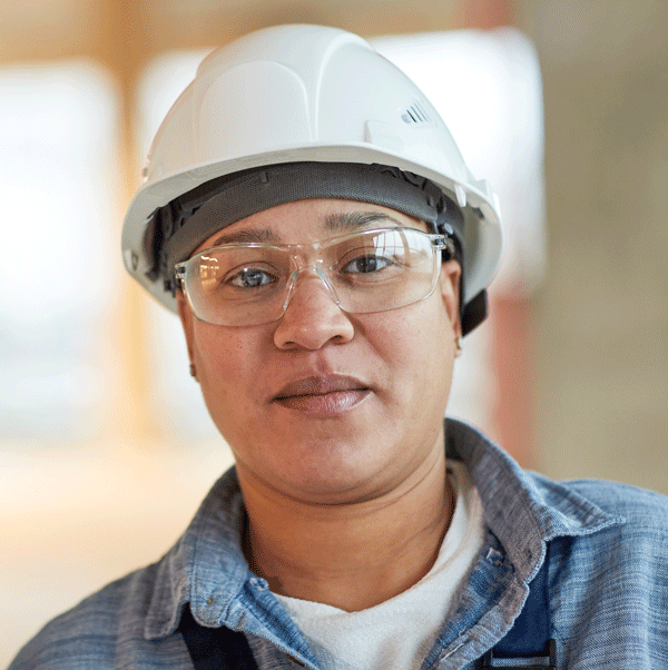 Woman in hard hat and safety glasses smiles, construction site background.