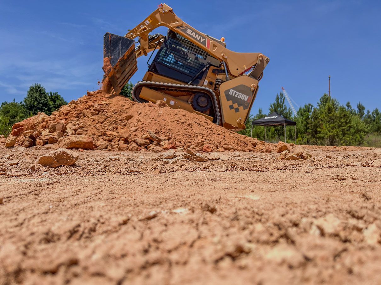 A yellow SANY compact track loader scoops red dirt on a sunny, outdoor construction site.