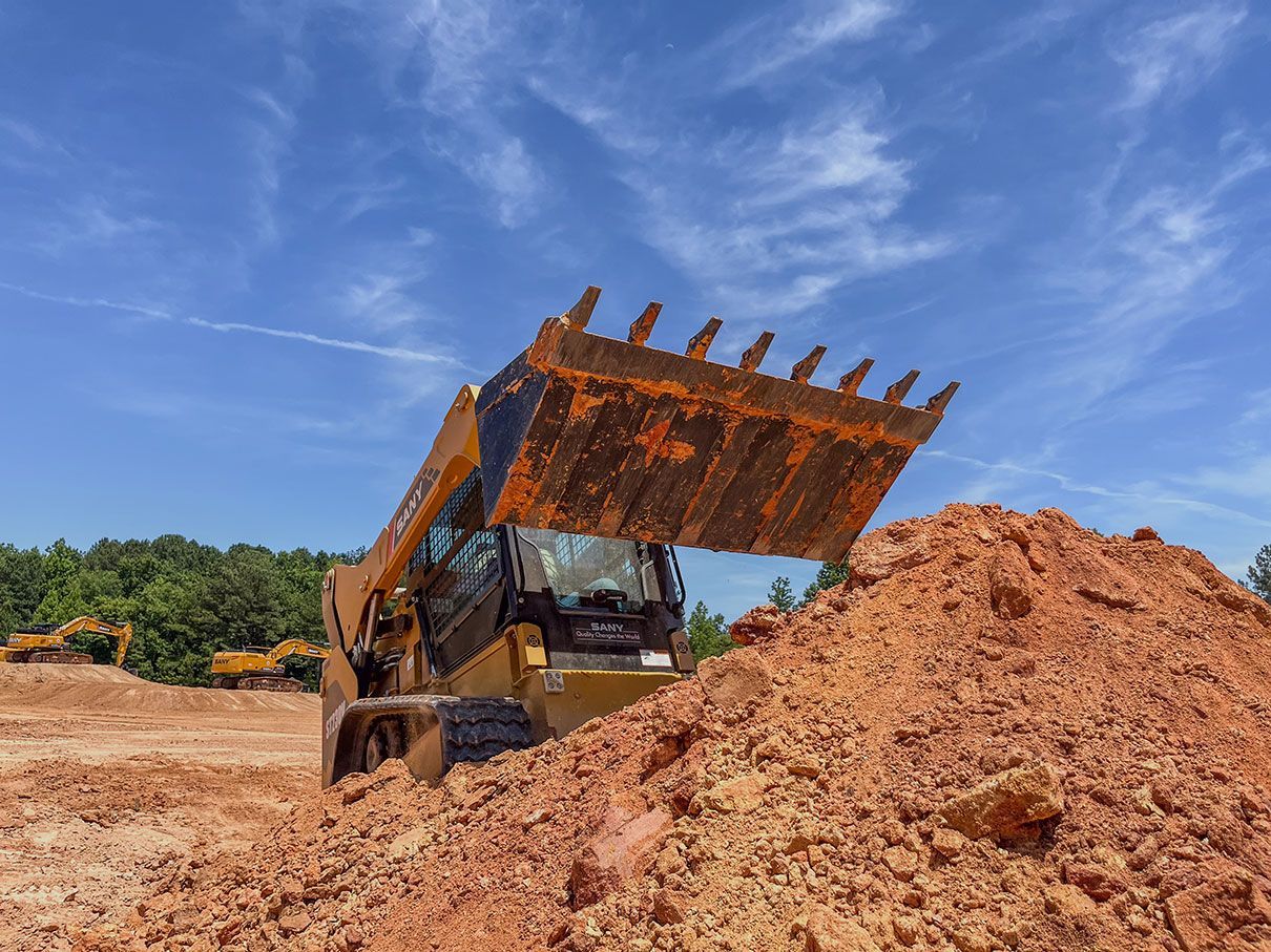 A yellow skid steer loader with a toothed bucket scoops red dirt at a construction site under a blue sky.