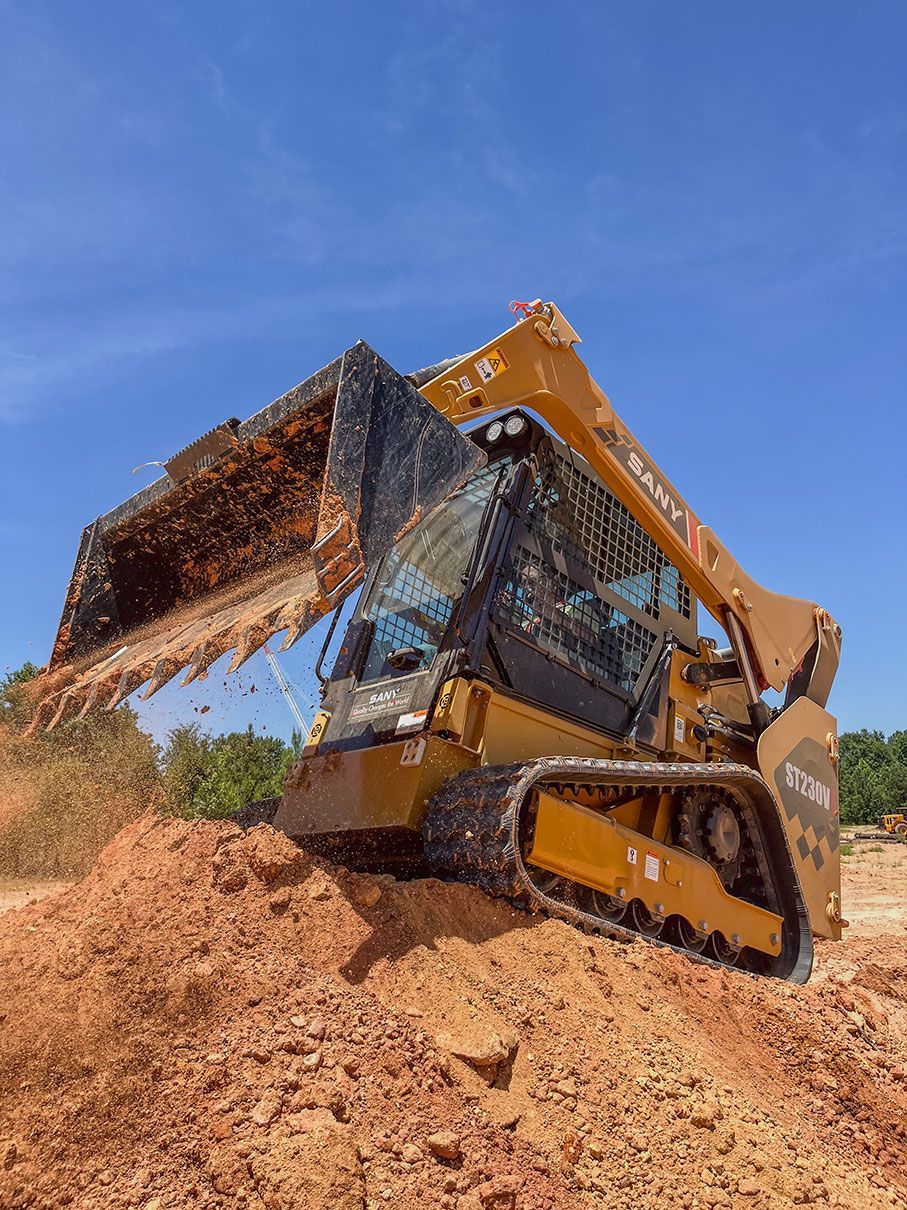 A yellow SANY skid steer loader with a bucket full of dirt climbs a large pile of earth against a blue sky.