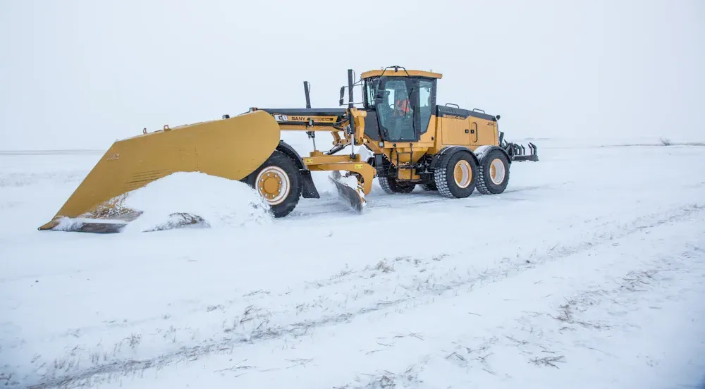 Yellow grader plowing snow on a flat, snow-covered surface.