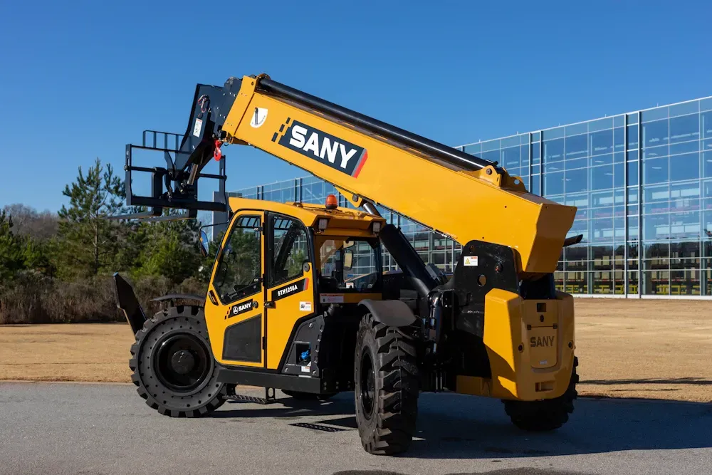A yellow SANY telehandler parked outdoors on a paved surface with a glass building in the background under a blue sky.