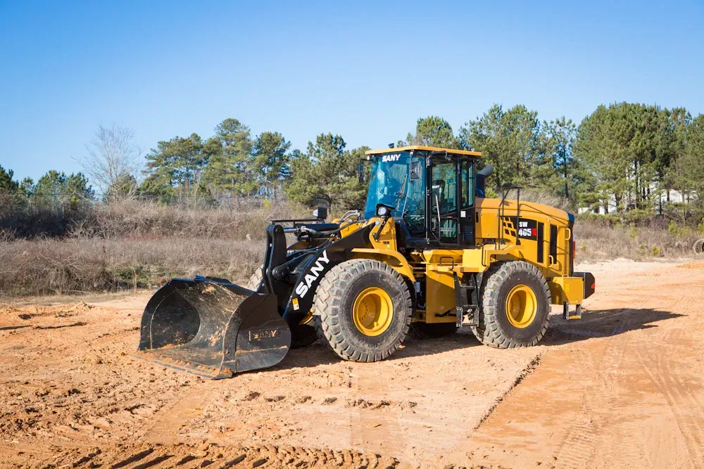 Yellow SANY wheel loader with bucket on a dirt road, blue sky, trees in the background.