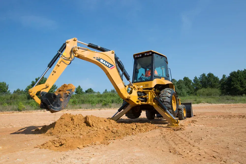 A yellow skid-steer loader dumping a pile of red soil at a construction site against a bright blue sky.