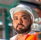 Man in hard hat, wearing orange safety vest, looking serious. Outdoors, likely construction site.