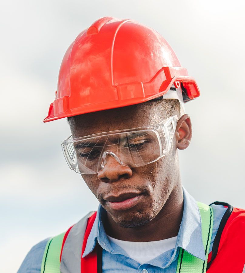Black construction worker in hard hat and safety glasses, looking down, sweat visible.