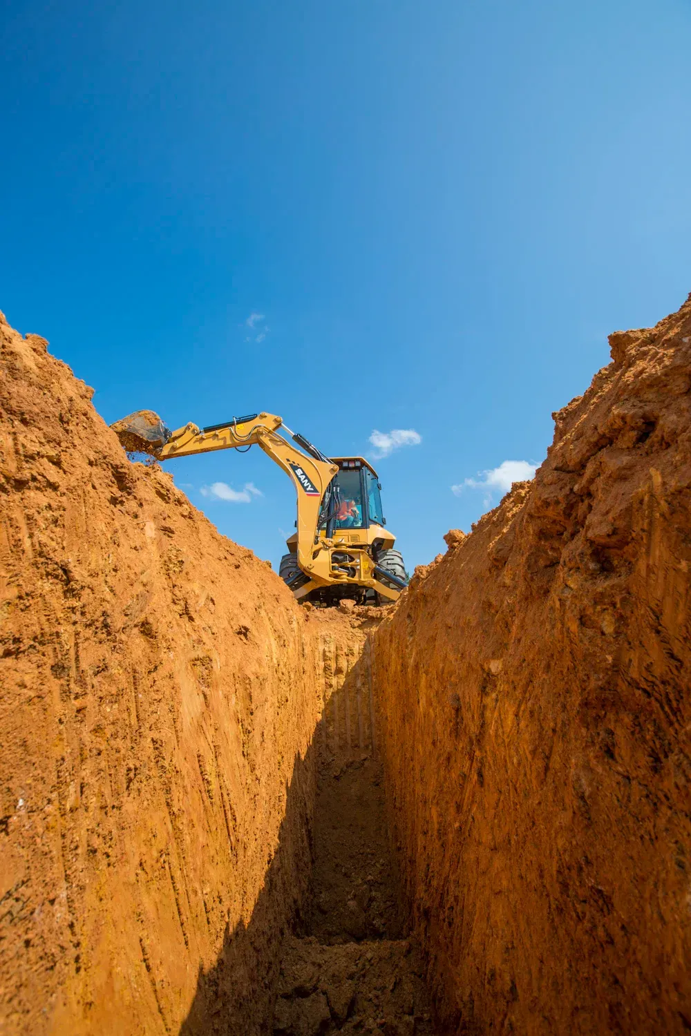 Yellow excavator digging a trench in reddish-brown soil under a bright blue sky.